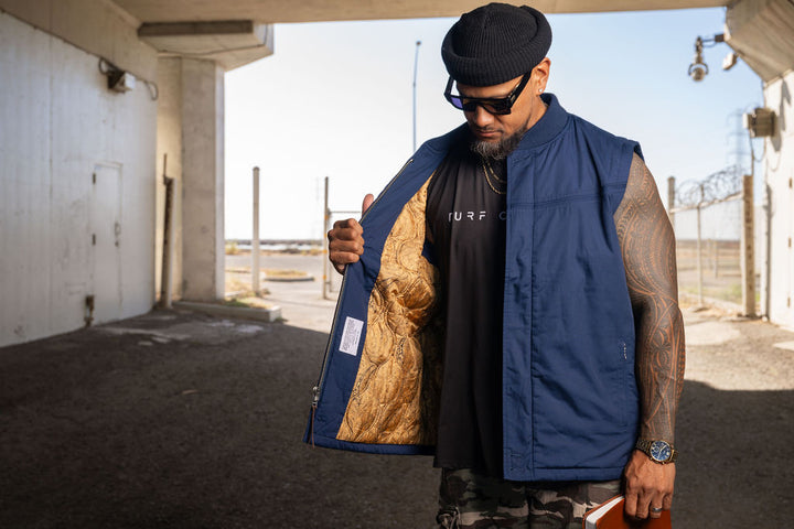 A man wearing a navy blue sleeveless vest over a black t-shirt stands in an outdoor concrete structure, holding the vest open to show a gold patterned lining.