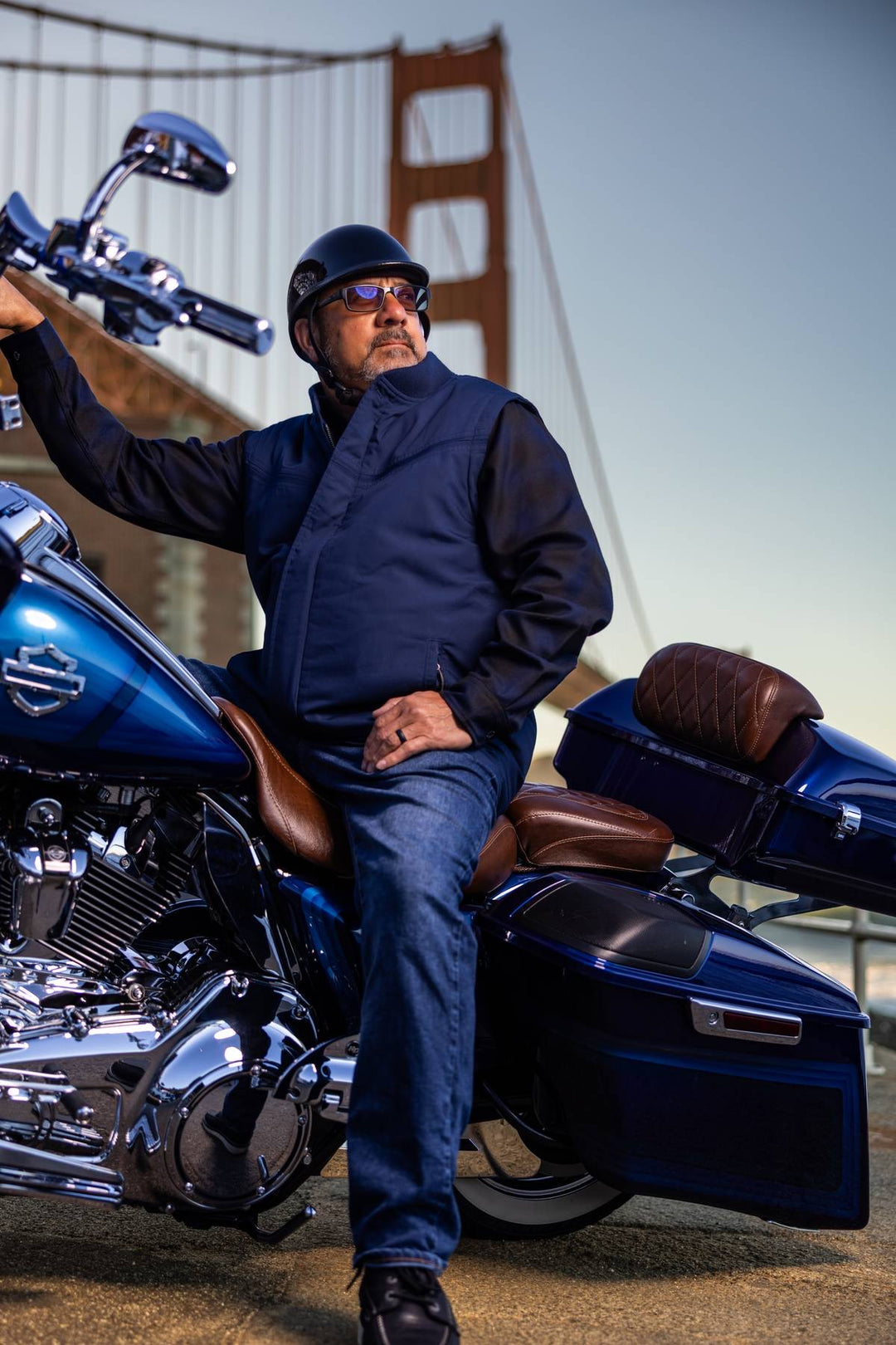 A man wearing a black helmet, sunglasses, and a dark navy jacket and jeans sits on a blue touring motorcycle in front of the Golden Gate Bridge.