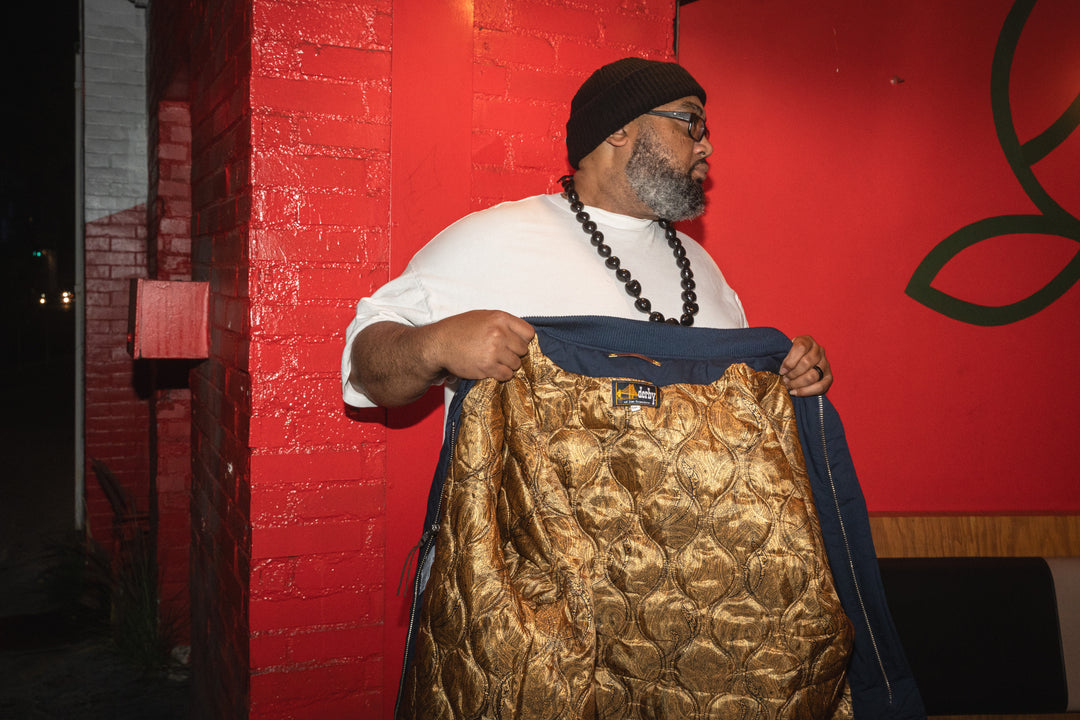 Man holding a gold patterned bag against a red wall with a logo.