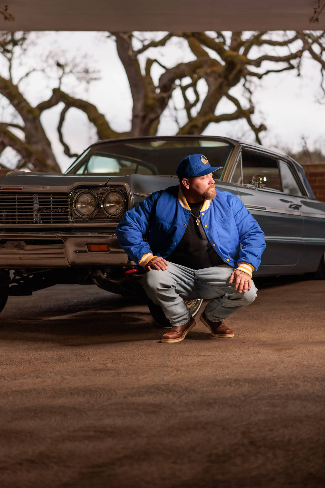 Man in blue jacket and cap squatting in front of a classic car with trees in the background