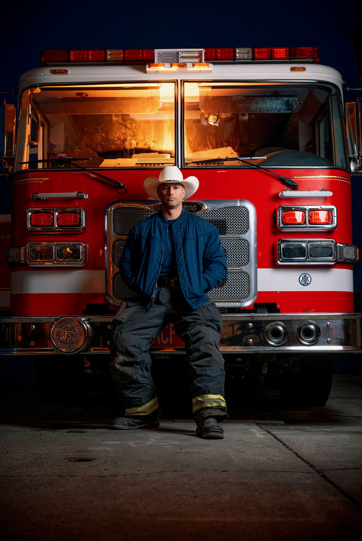 A person wearing a navy blue jacket with a gold paisley pattern, sitting in front of a fire engine.