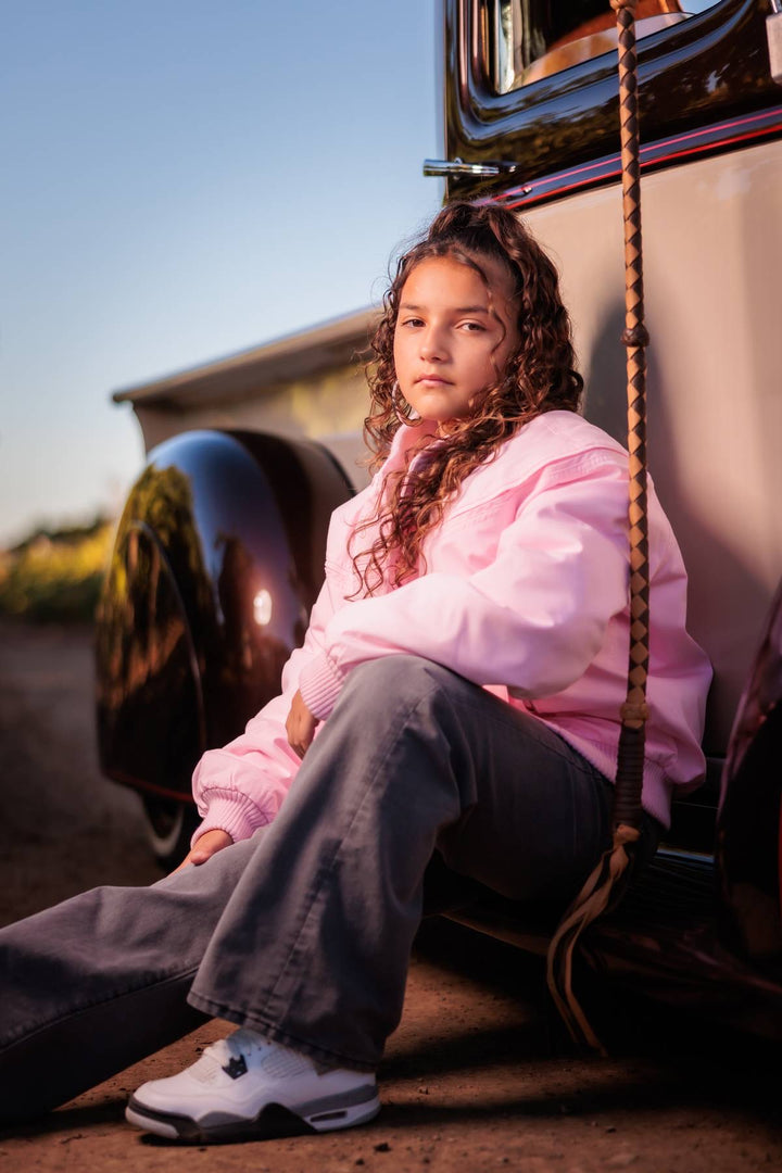 A young girl with curly brown hair sits on the bumper of a vintage car outdoors, wearing a light pink zip-up hoodie, dark gray wide-leg jeans, and white sneakers.