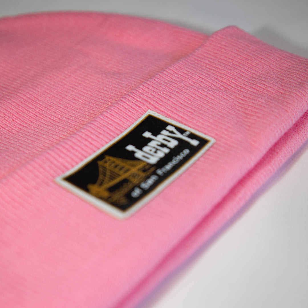 A close-up of a pink ribbed knit beanie with a black rectangular patch that reads 'derby of San Francisco' and shows a gold bridge graphic.