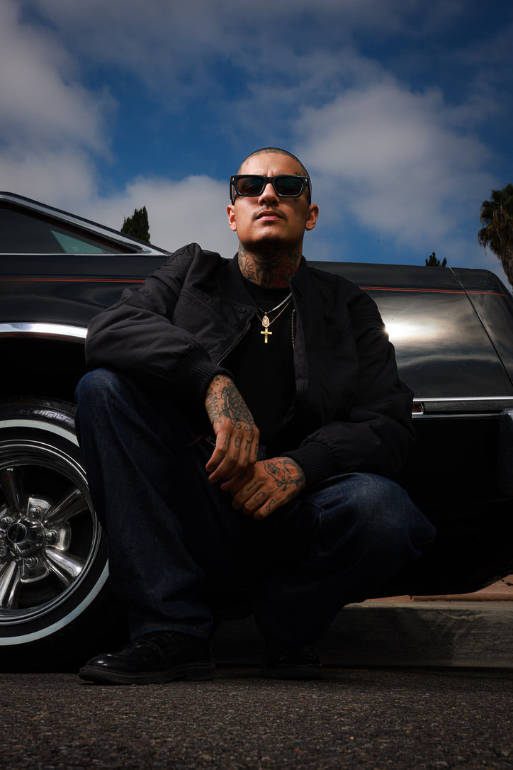 A tattooed man wearing black sunglasses, a black jacket, and dark jeans crouches in front of a black classic car under a blue sky with clouds.