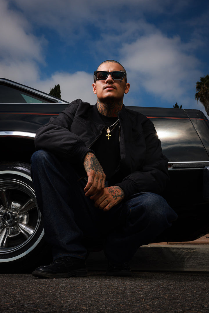 A tattooed man wearing black sunglasses, a black jacket, and dark jeans crouches in front of a black classic car under a blue sky with clouds.