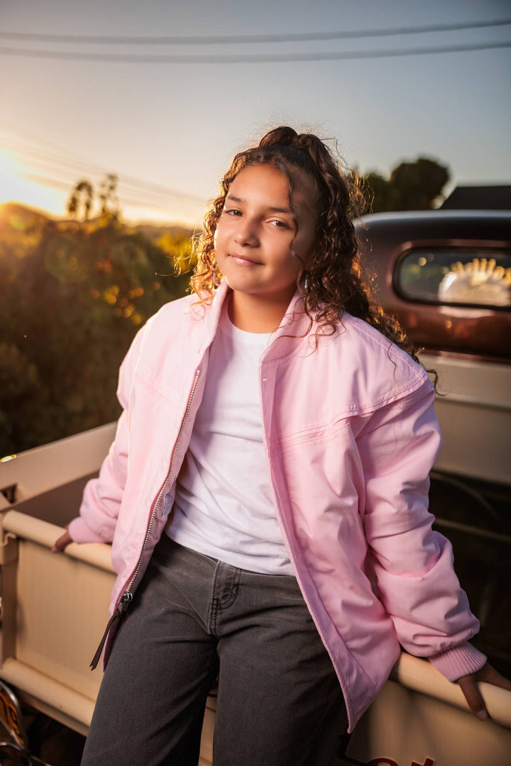 A young woman with curly brown hair stands in the bed of a vintage pickup truck at sunset, wearing a light pink bomber jacket over a white t-shirt and dark gray jeans.