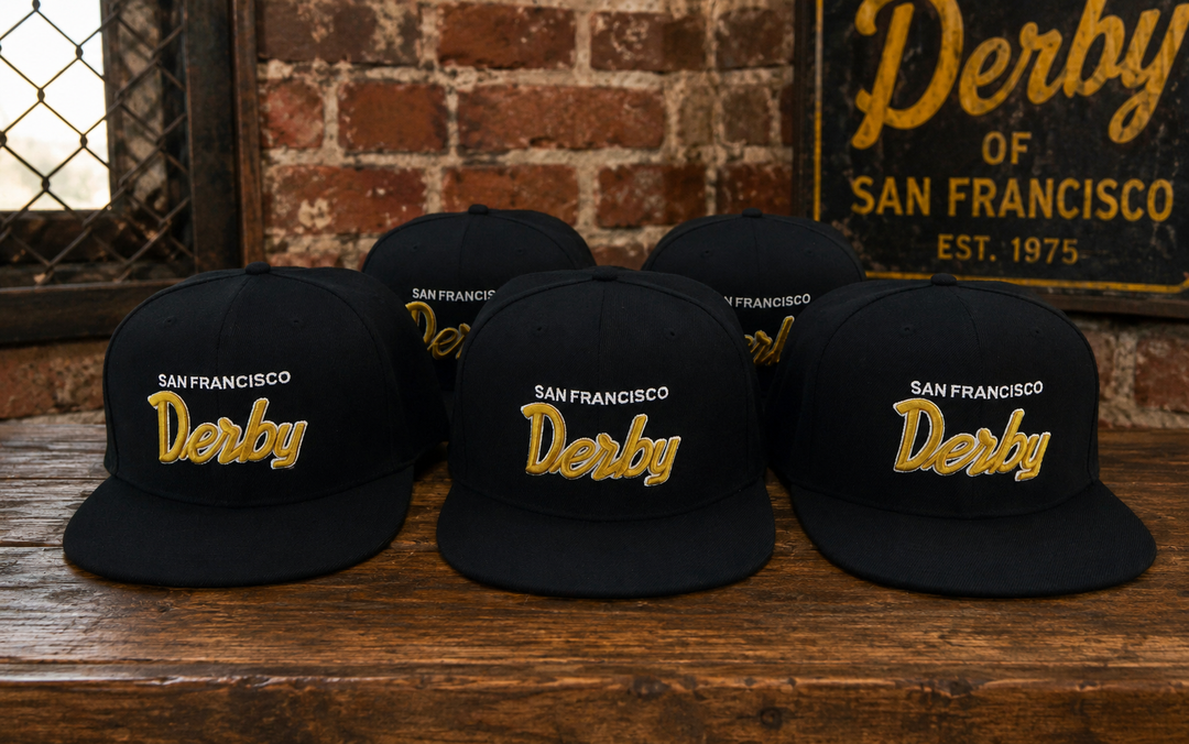 Five black San Francisco Derby snapback caps arranged on a rustic wooden table in front of a brick wall, with a vintage-style Derby sign in the background.