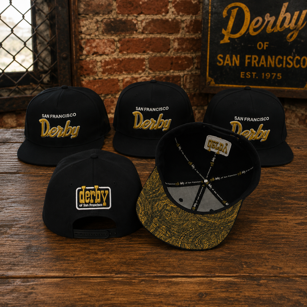 Five black snapback caps with 'San Francisco Derby' branding in gold and white, arranged on a rustic wooden table with a brick wall and vintage sign in the background.