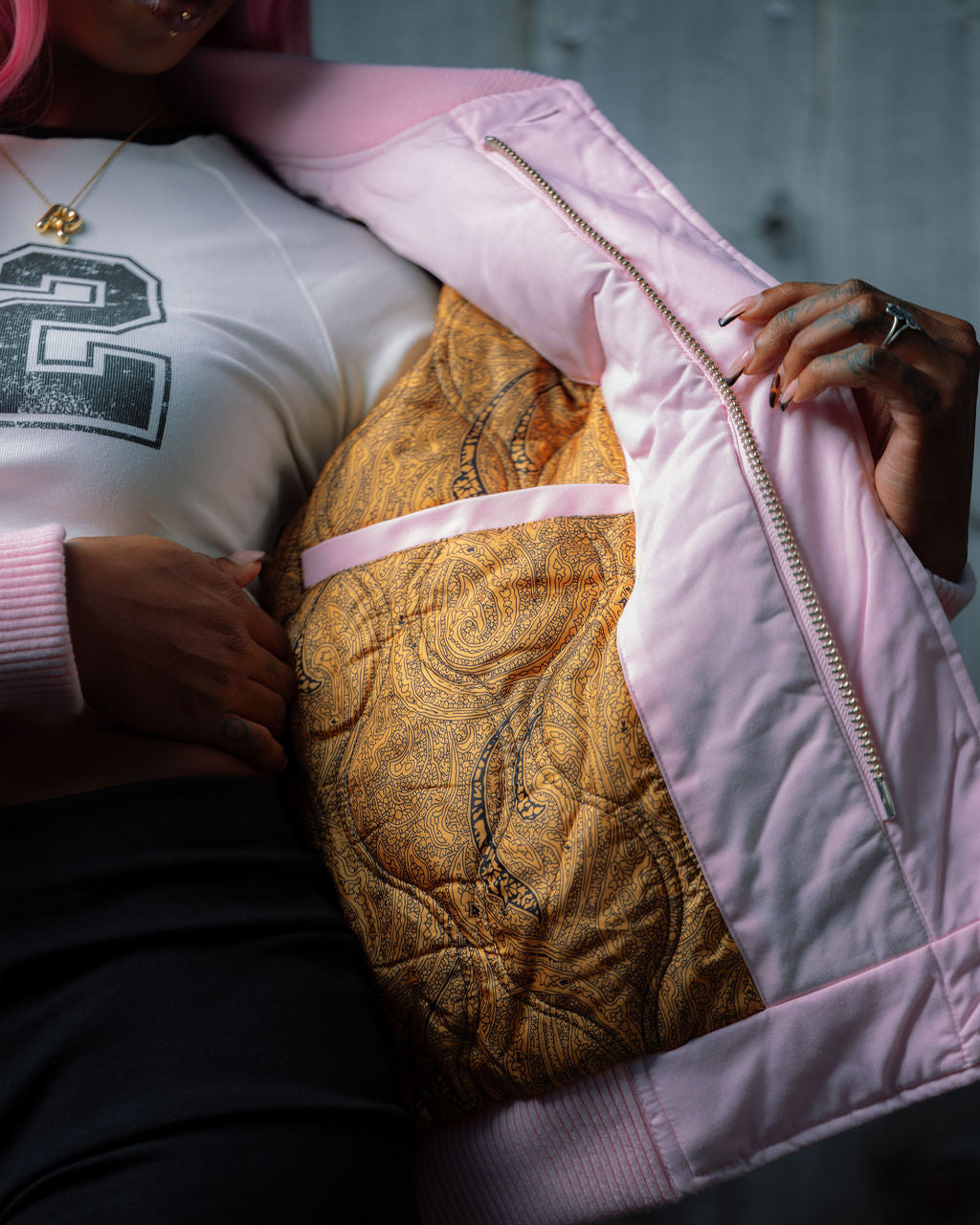 A close-up lifestyle shot of a person wearing a light pink quilted bomber jacket, unzipped to reveal a gold patterned quilted lining. The person is also wearing a white graphic t-shirt and black pants.