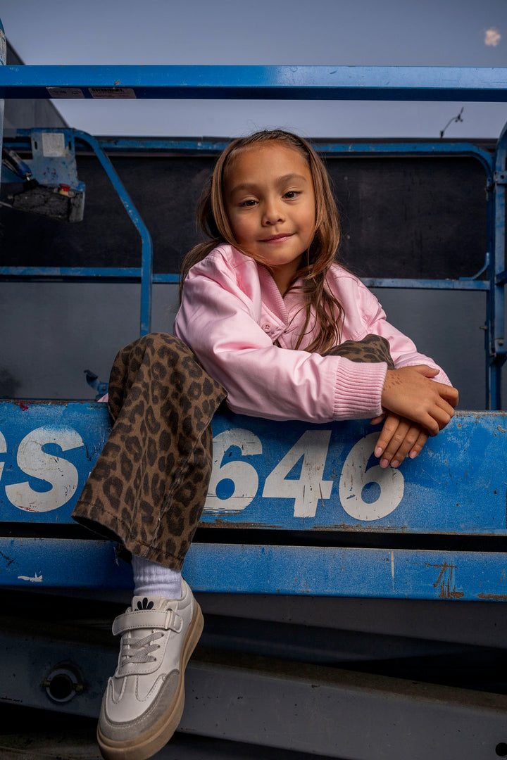 A young girl with long brown hair sits on the blue metal bed of a truck, wearing a pink zip-up hoodie, leopard-print pants, white socks, and white sneakers.