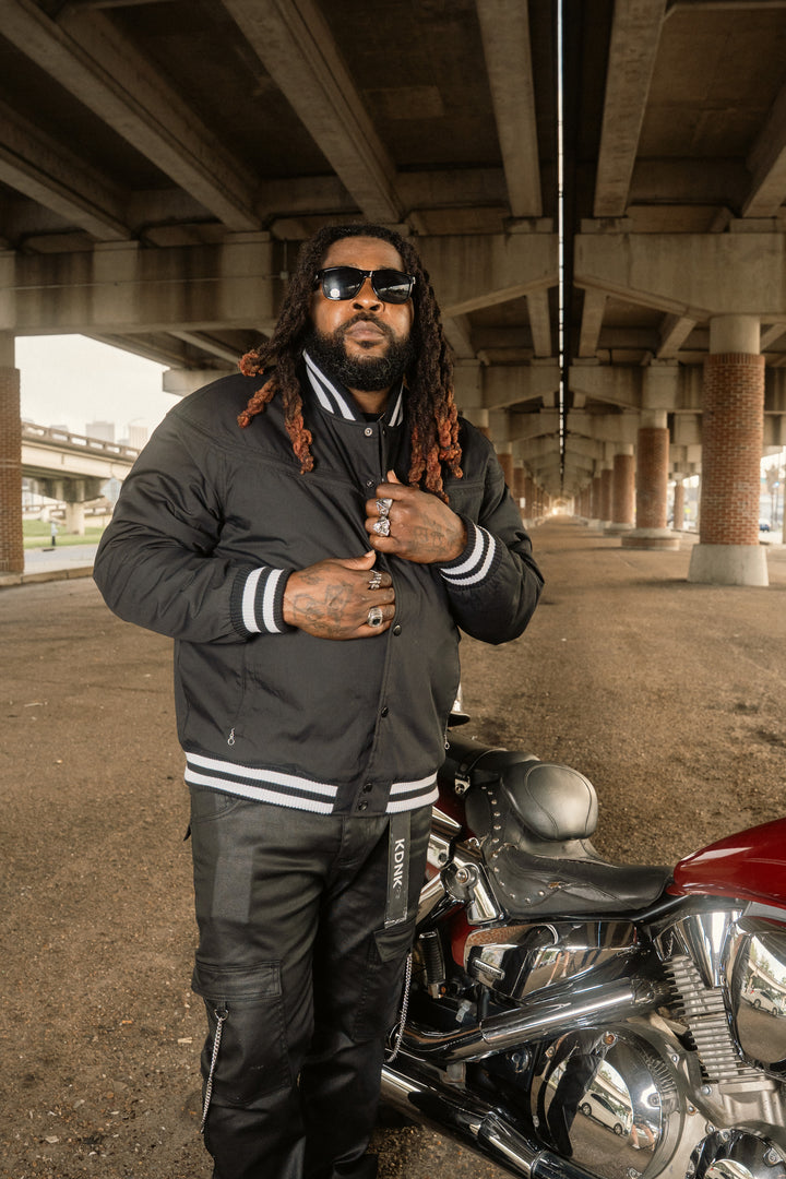 Man with dreadlocks wearing sunglasses and a black jacket standing next to a motorcycle under an overpass.