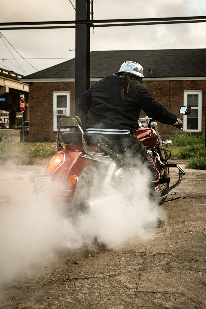 Person riding a motorcycle with dust in the foreground, brick building in the background