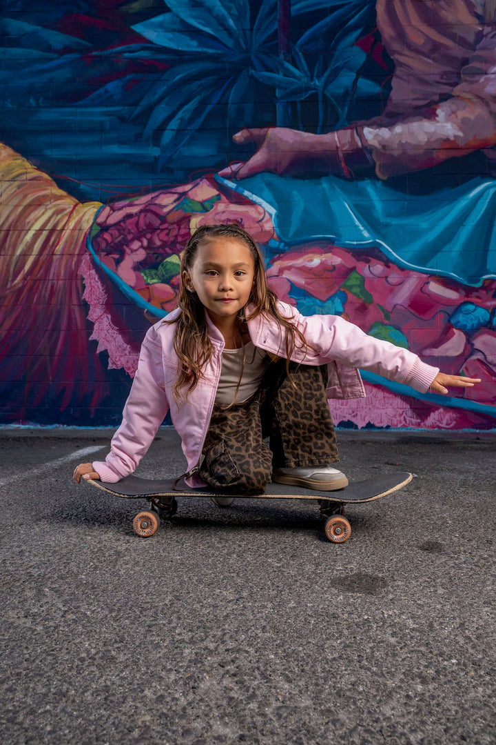 A young girl wearing a light pink zip-up jacket, leopard-print pants, and gray sneakers is balancing on a skateboard in front of a colorful mural.