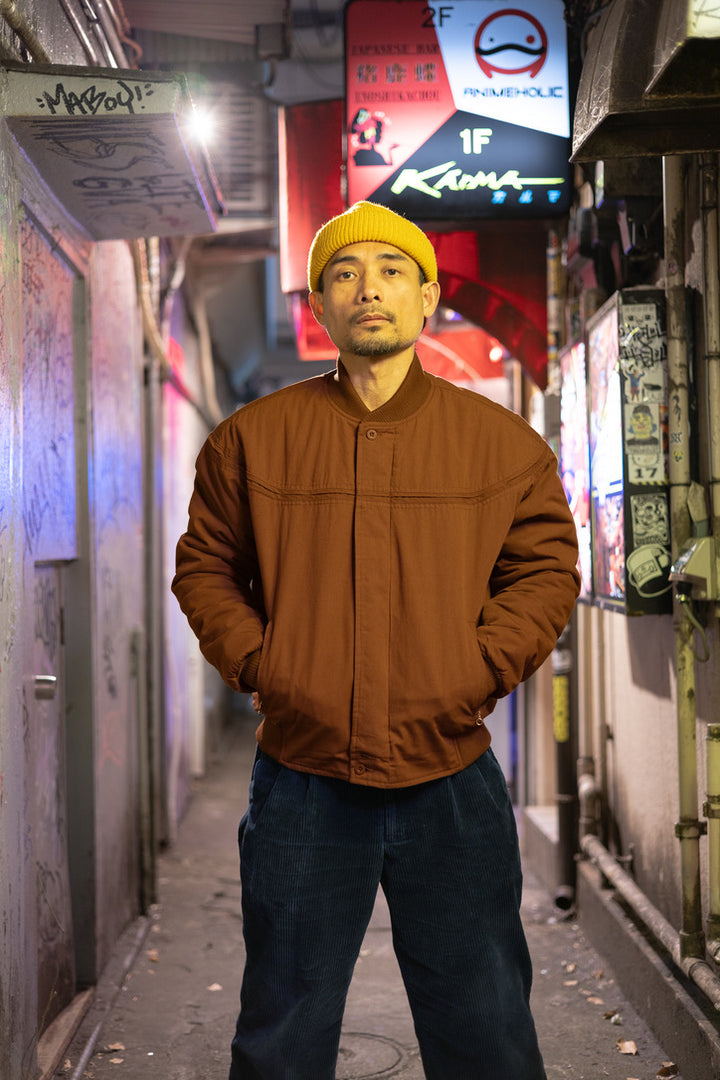 Man wearing a brown jacket and yellow beanie standing in an urban alleyway with signs in the background.