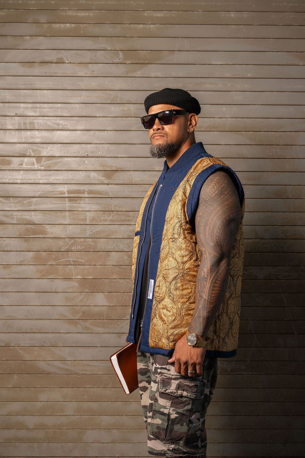 A man wearing a gold patterned vest over a blue shirt, camouflage pants, and a black beret, standing against a brown wood-paneled wall.