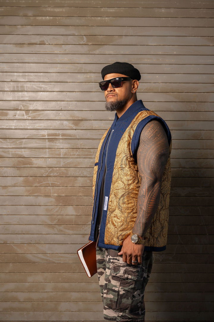 A man wearing a gold patterned vest over a blue shirt, camouflage pants, and a black beret, standing against a brown wood-paneled wall.