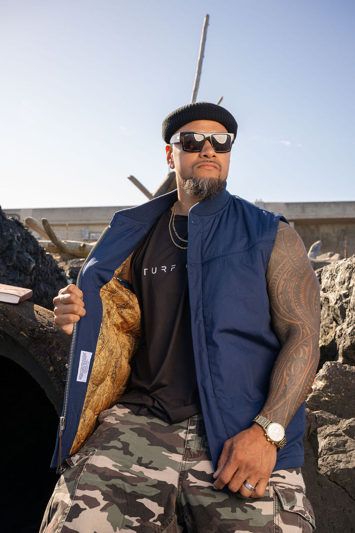 A man wearing a navy blue zippered vest over a black t-shirt, camouflage pants, and a black beanie, sitting outdoors in front of a stone wall under a clear blue sky.