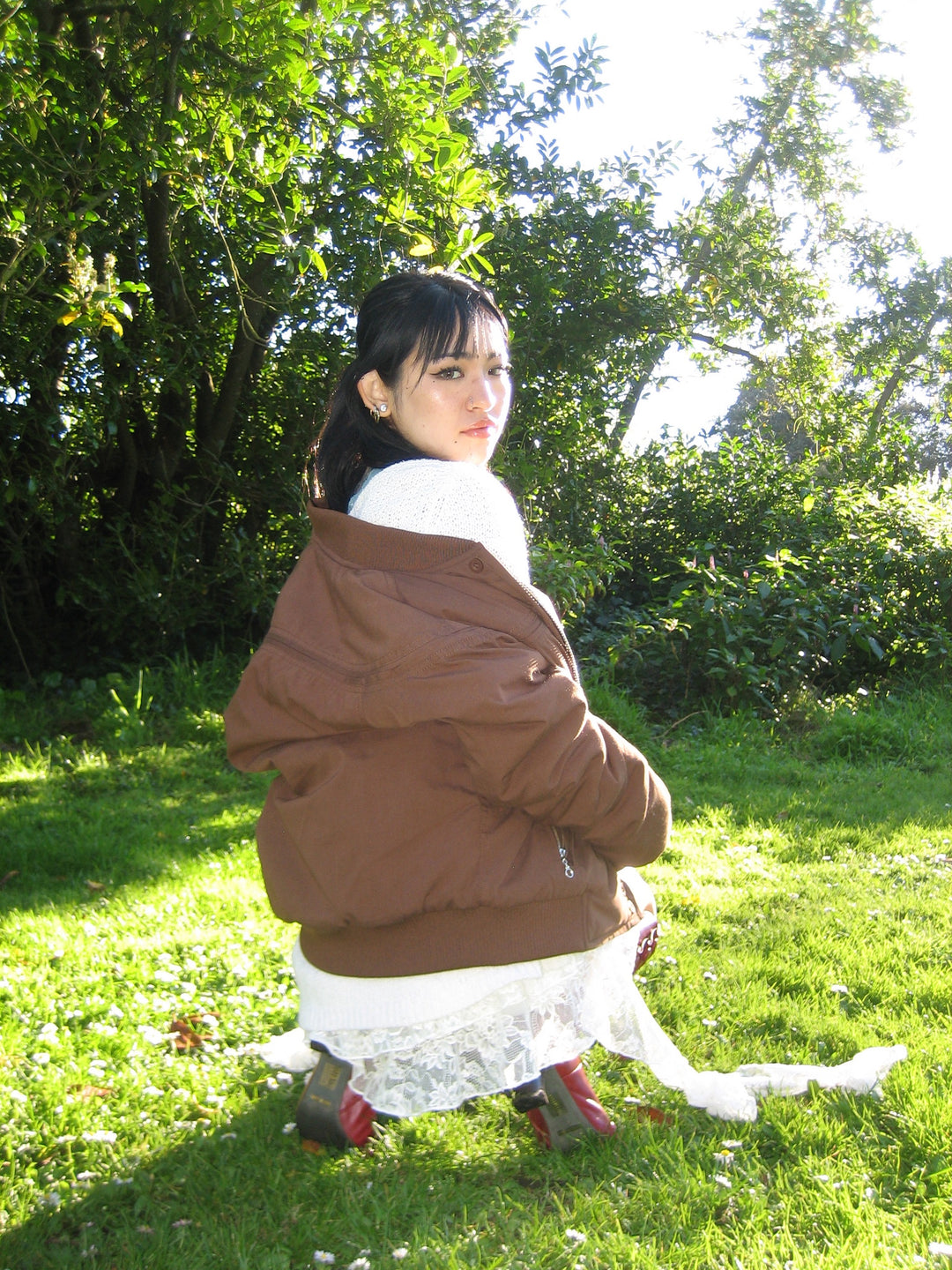 Woman in a brown coat and white dress standing in a grassy area with trees in the background