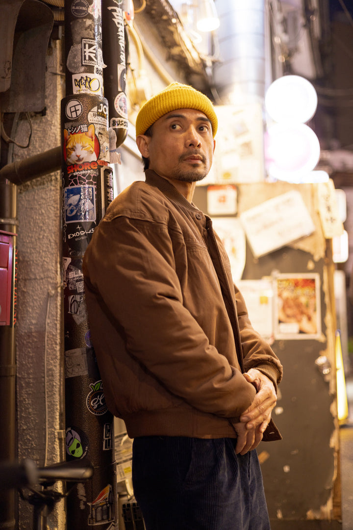 Man wearing a brown jacket and yellow beanie standing in an urban alleyway with posters on the wall.