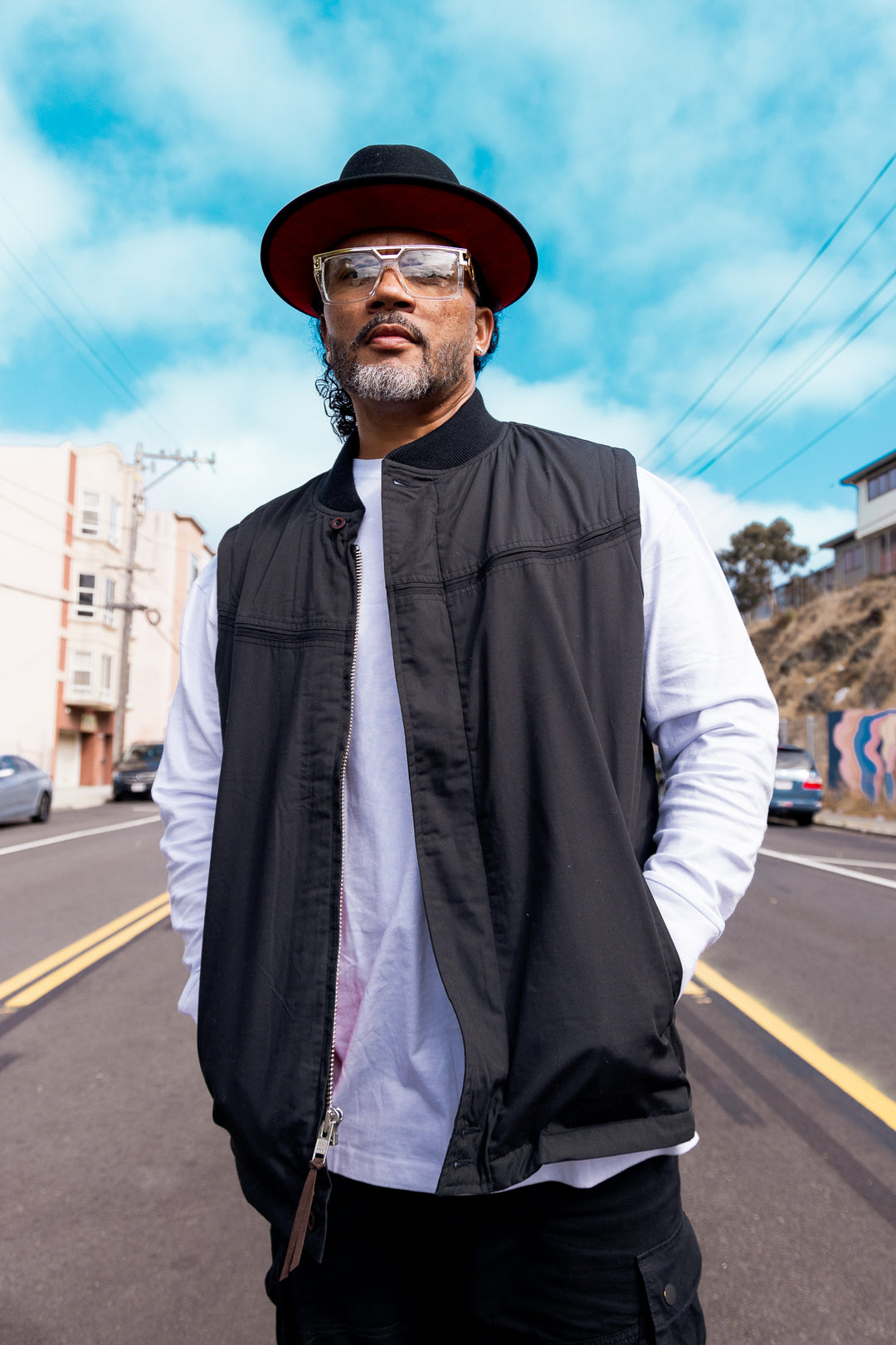 Man wearing a black vest, white shirt, and hat on a street with buildings and blue sky in the background