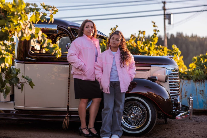 Two women standing outdoors next to a vintage cream and dark brown car, wearing matching light pink jackets. The scene is set in a vineyard with grapevines and warm golden sunlight.
