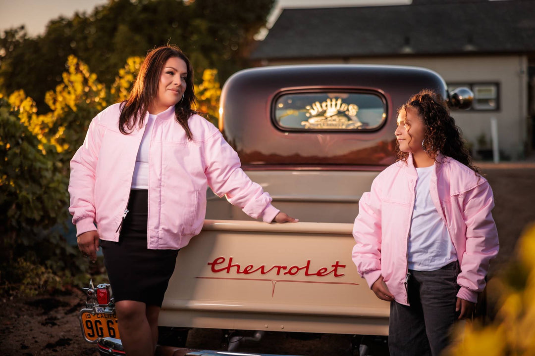 Two women wearing matching light pink bomber jackets stand in front of a vintage Chevrolet pickup truck outdoors at sunset. One woman leans on the truck bed while the other stands beside her, with trees and a house visible in the background.