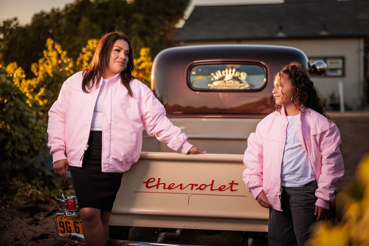 Two women wearing matching light pink bomber jackets stand in front of a vintage Chevrolet pickup truck outdoors at sunset. One woman leans on the truck bed while the other stands beside her, with trees and a house visible in the background.