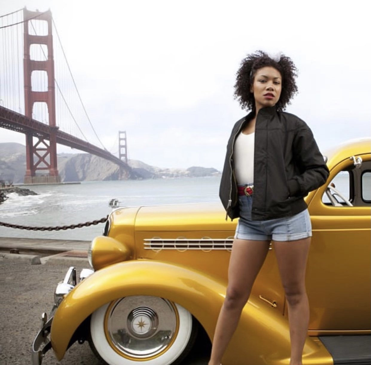 Young woman posing in front of a yellow old school car wearing a Black Classic Derby Jacket